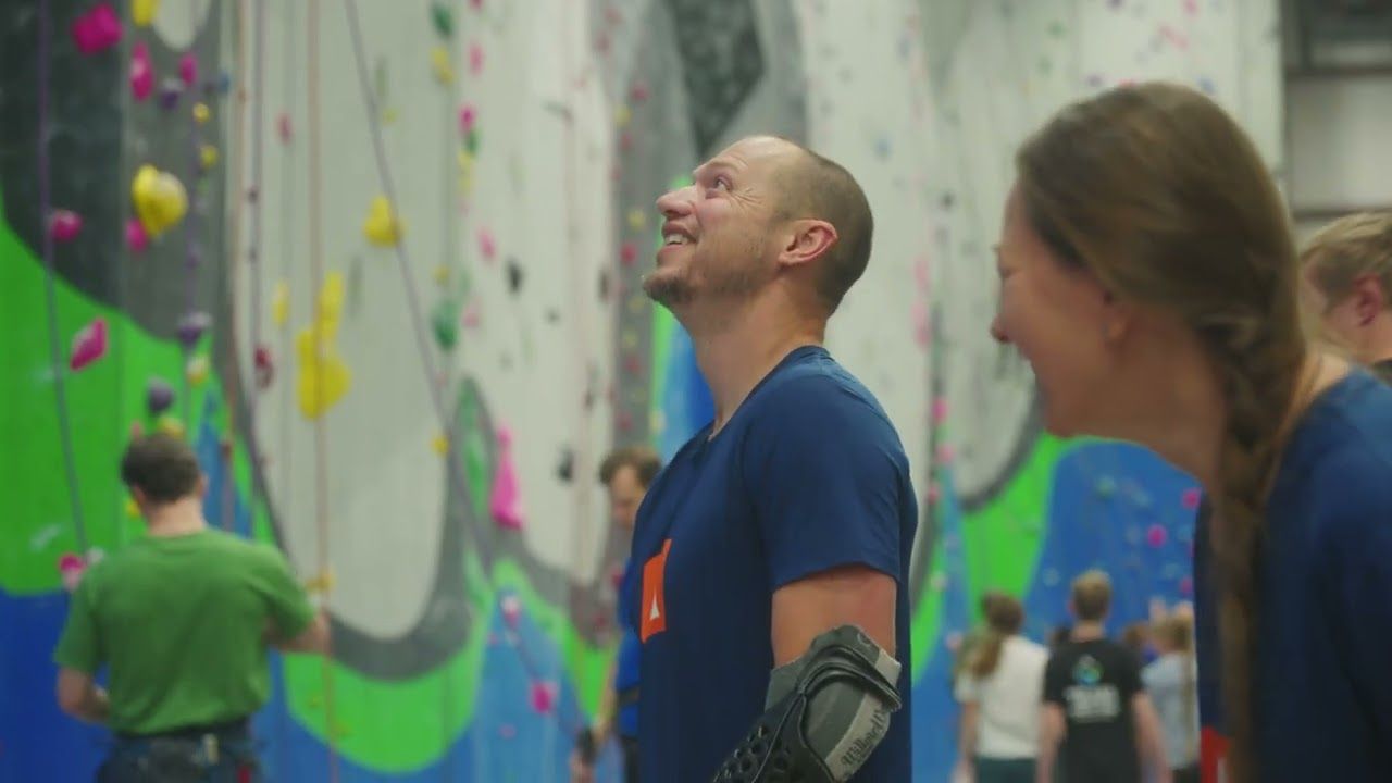 A man smiling as he looks up at an indoor climbing wall, wearing an adaptive climbing prosthesis on his forearm