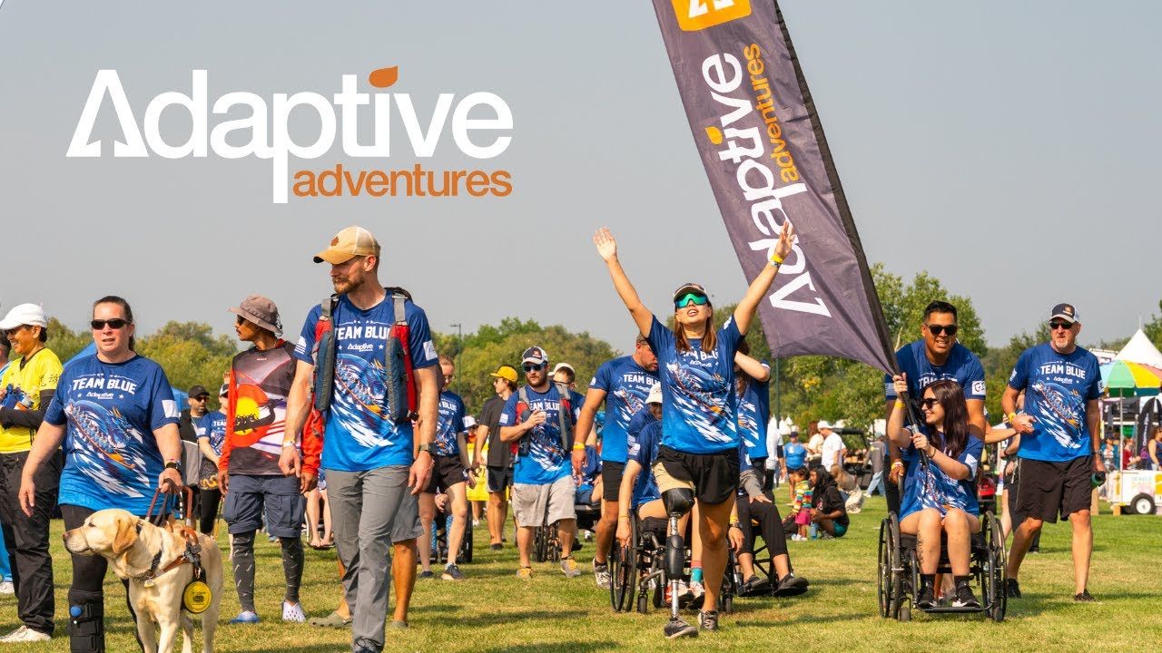 Adaptive Adventures participants at an outdoor event in team jerseys, one walking toward the camera on a running prosthesis with both arms raised