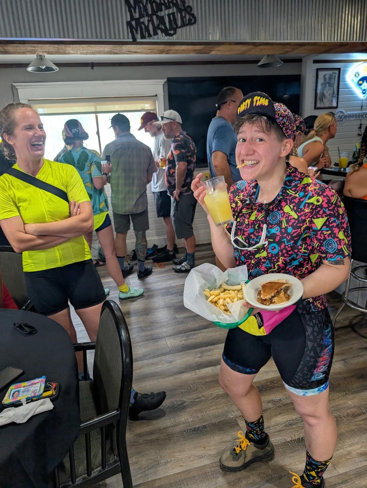A rider in a brightly patterned kit grinning with a plate of food and a drink inside a bar