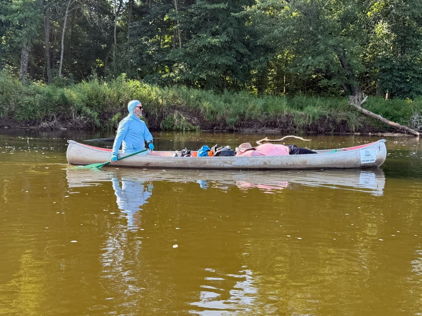 A lone paddler in a loaded canoe on a still, green stretch of river