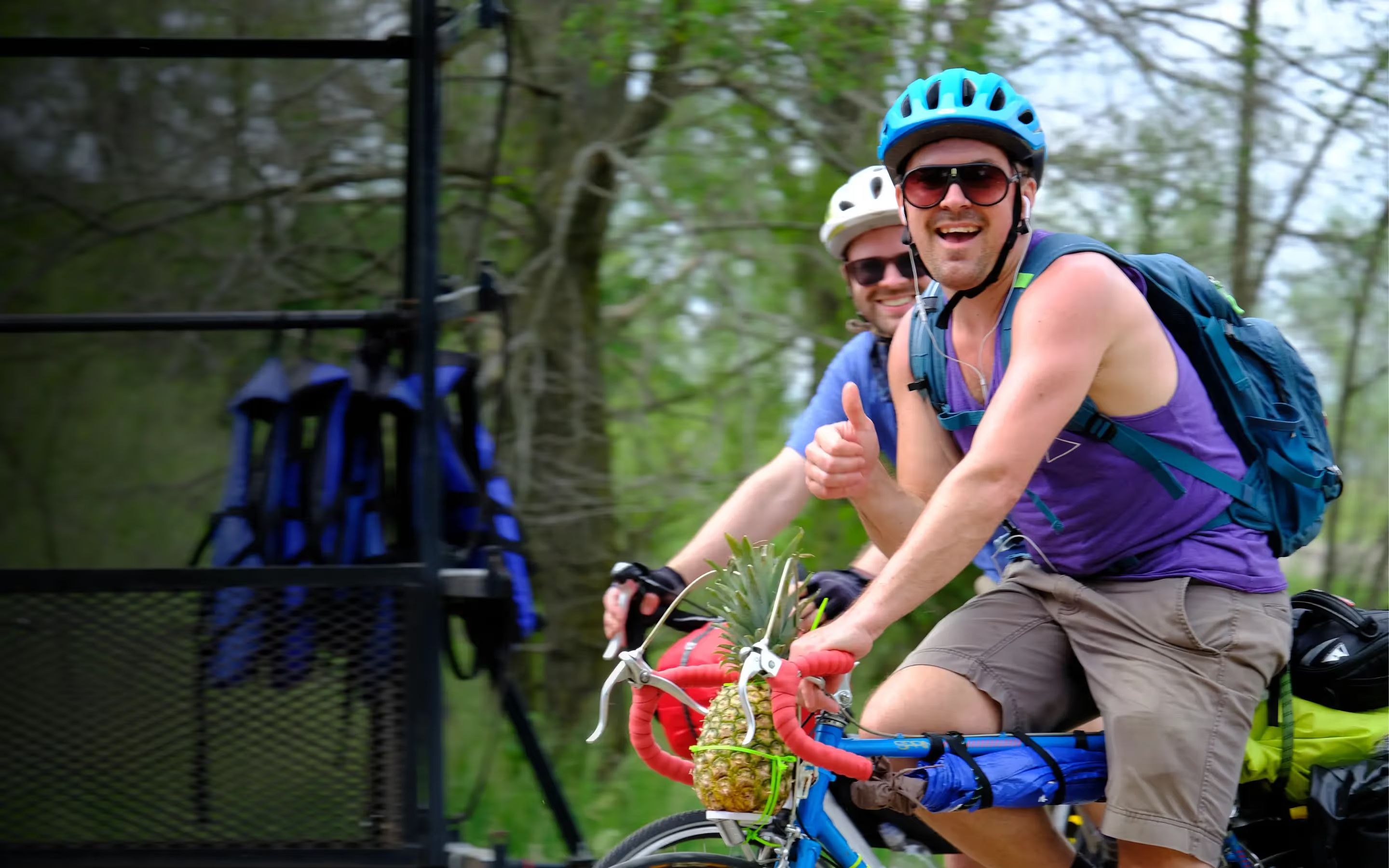 Two riders grinning on loaded bikes, one with a pineapple strapped to the handlebars