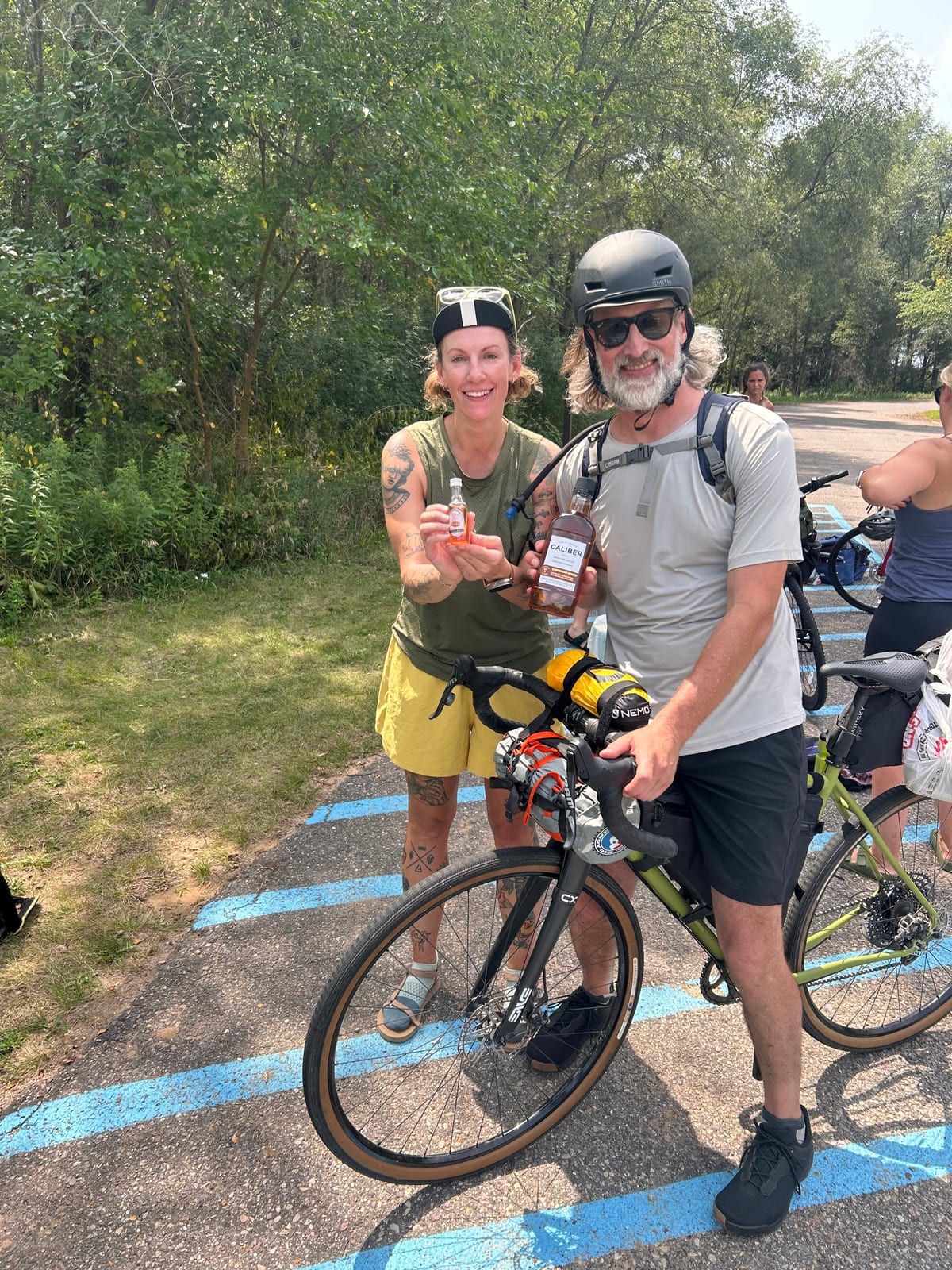 Two riders standing with their bikes on a paved path, holding drinks and snacks