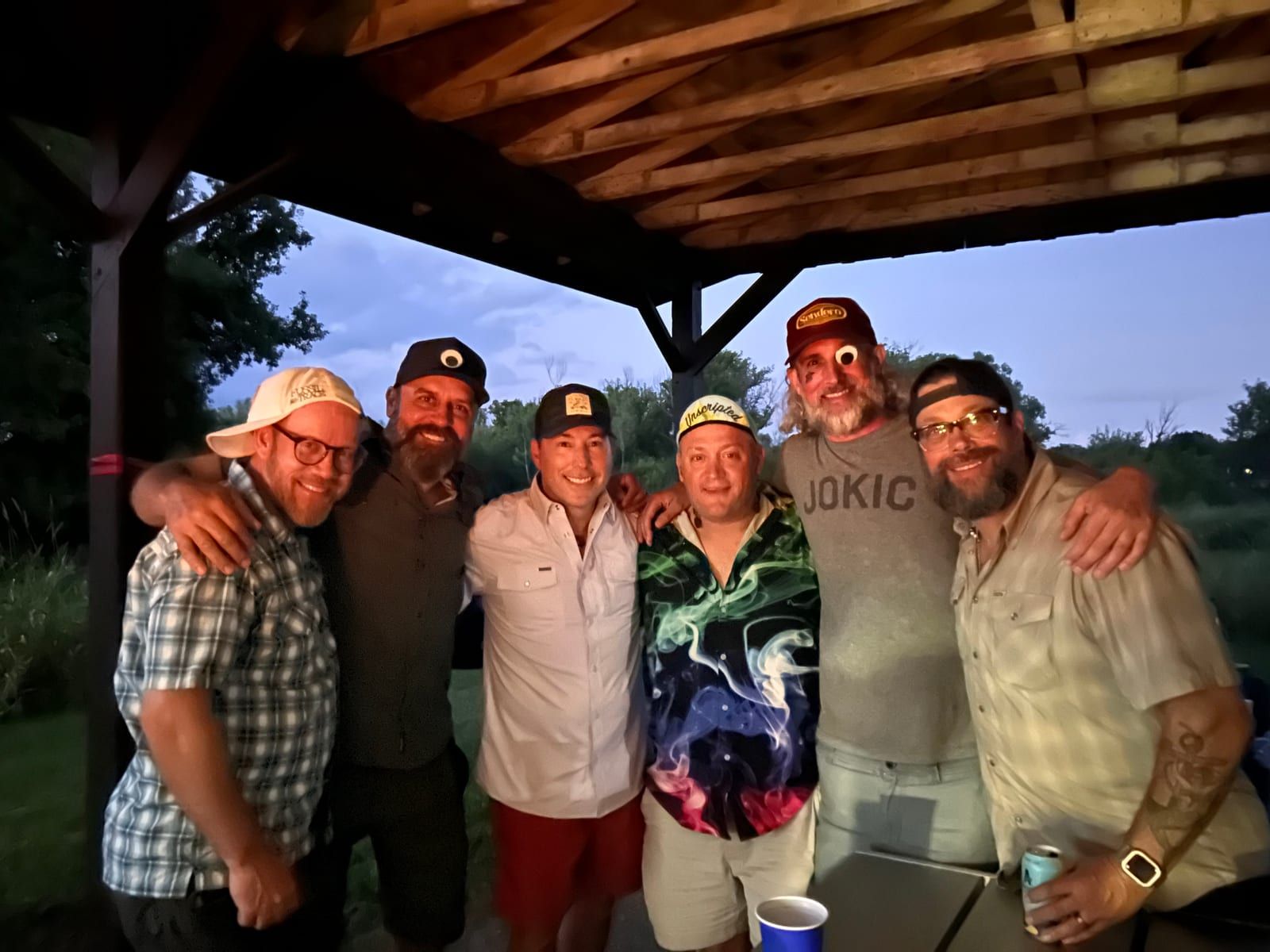 Six riders with their arms around each other under a wooden shelter at dusk