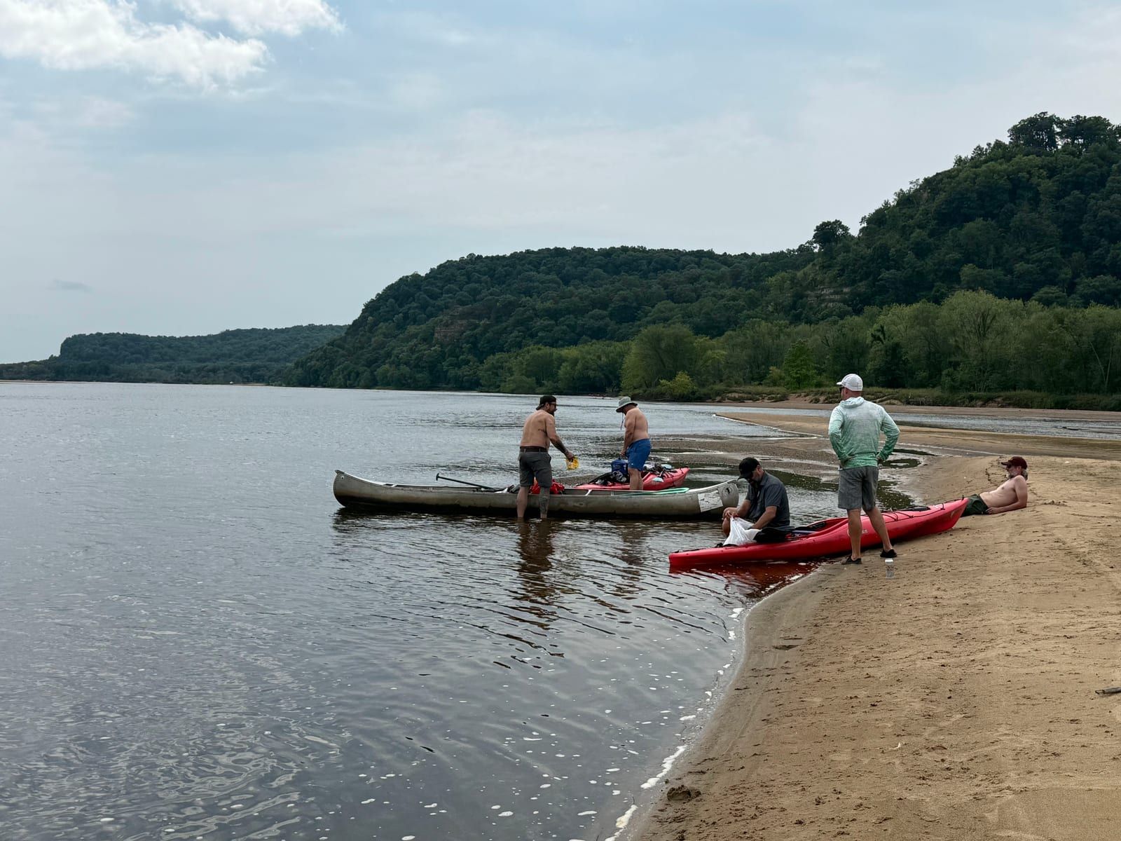 Canoes and kayaks pulled up on a wide sandbar with wooded bluffs across the river
