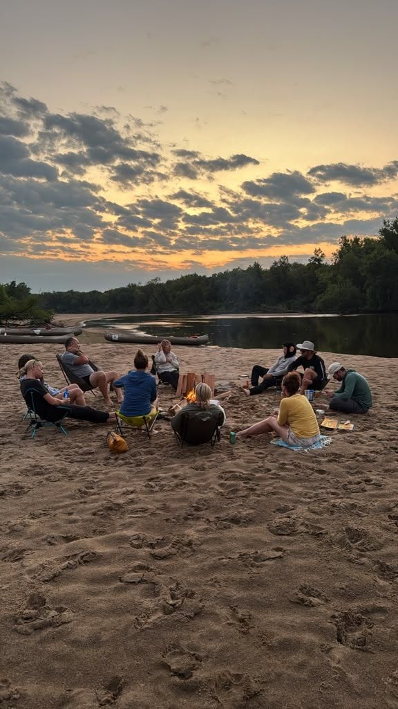 A group sitting on a river sandbar at dusk with kayaks pulled up on the sand