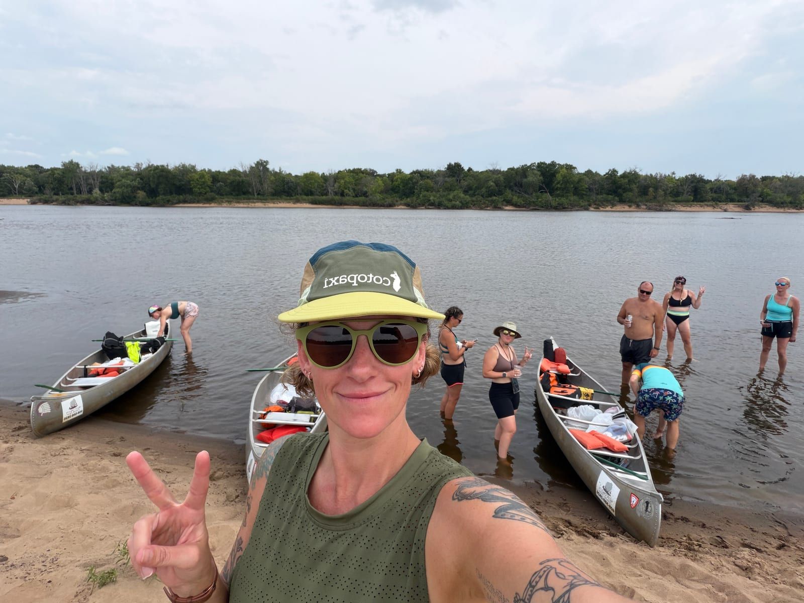A paddler throwing a peace sign in front of beached canoes while others wade in the shallows