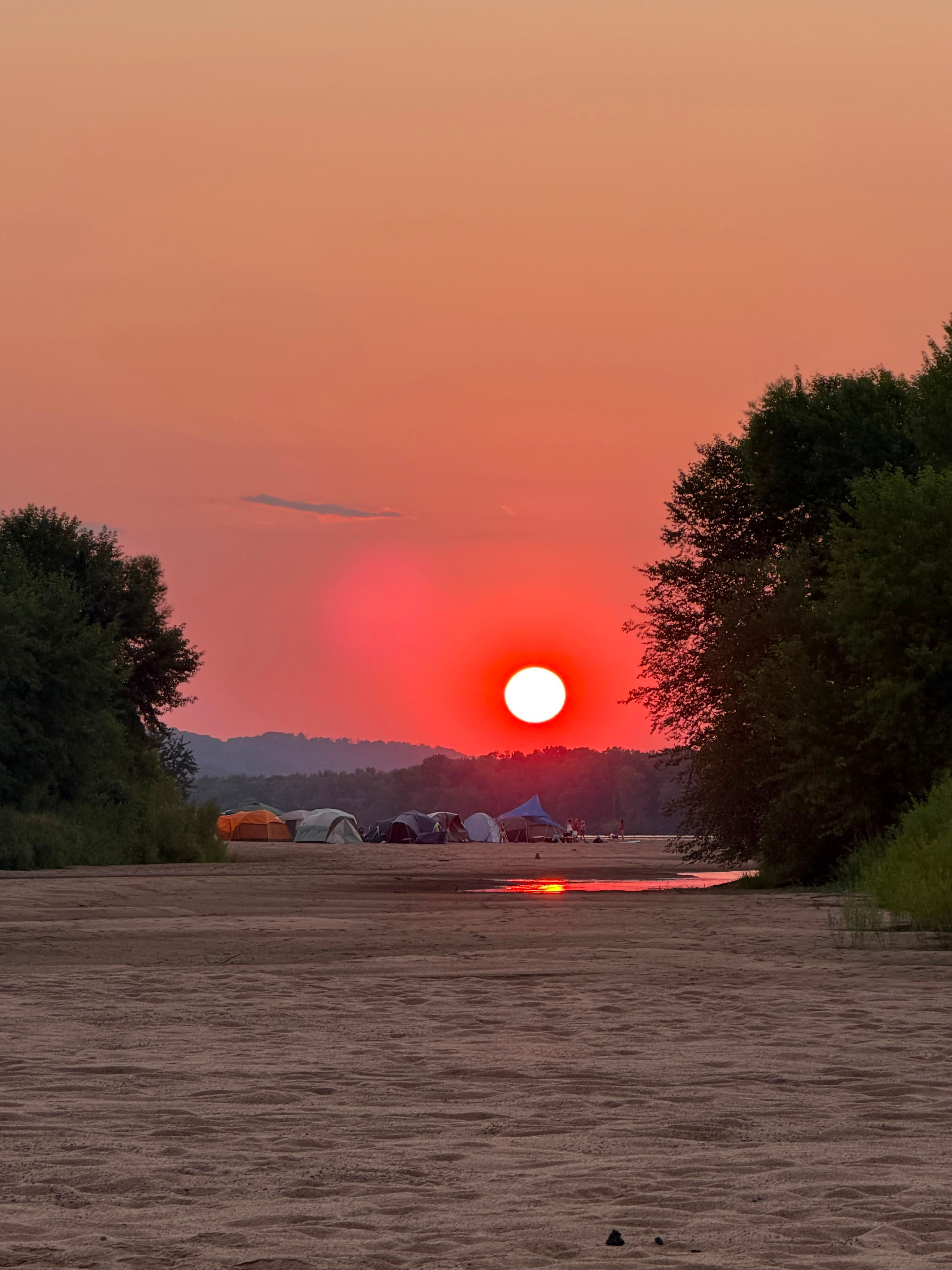 A red sun setting over a river sandbar with tents pitched along the shore