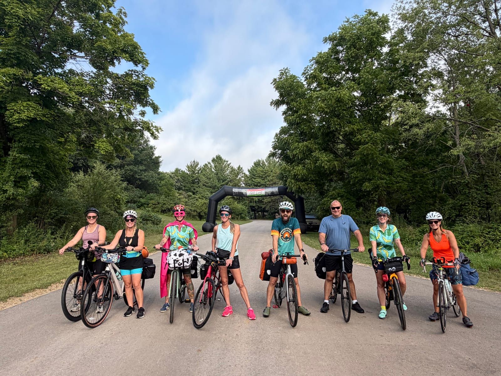 Nine riders lined up with loaded bikes on a country road beneath the inflatable start arch