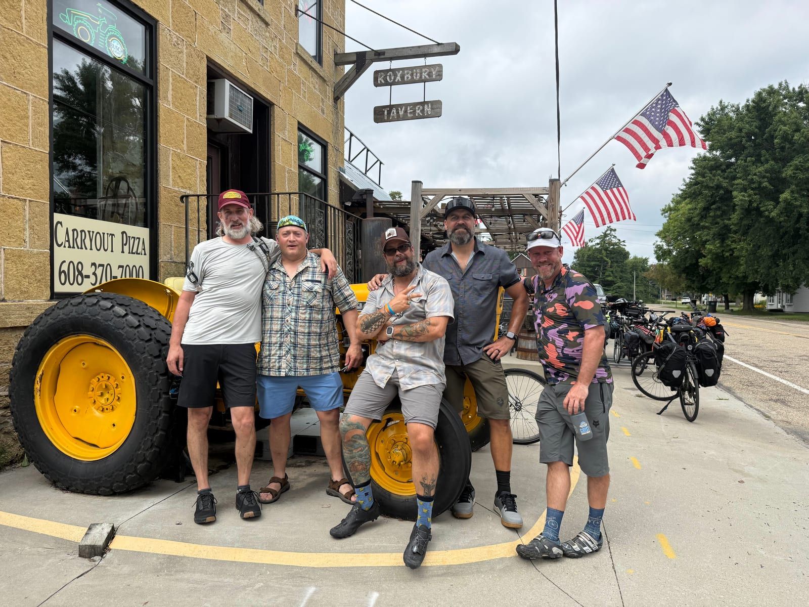 Six riders posing on and around a yellow tractor parked outside a small-town tavern